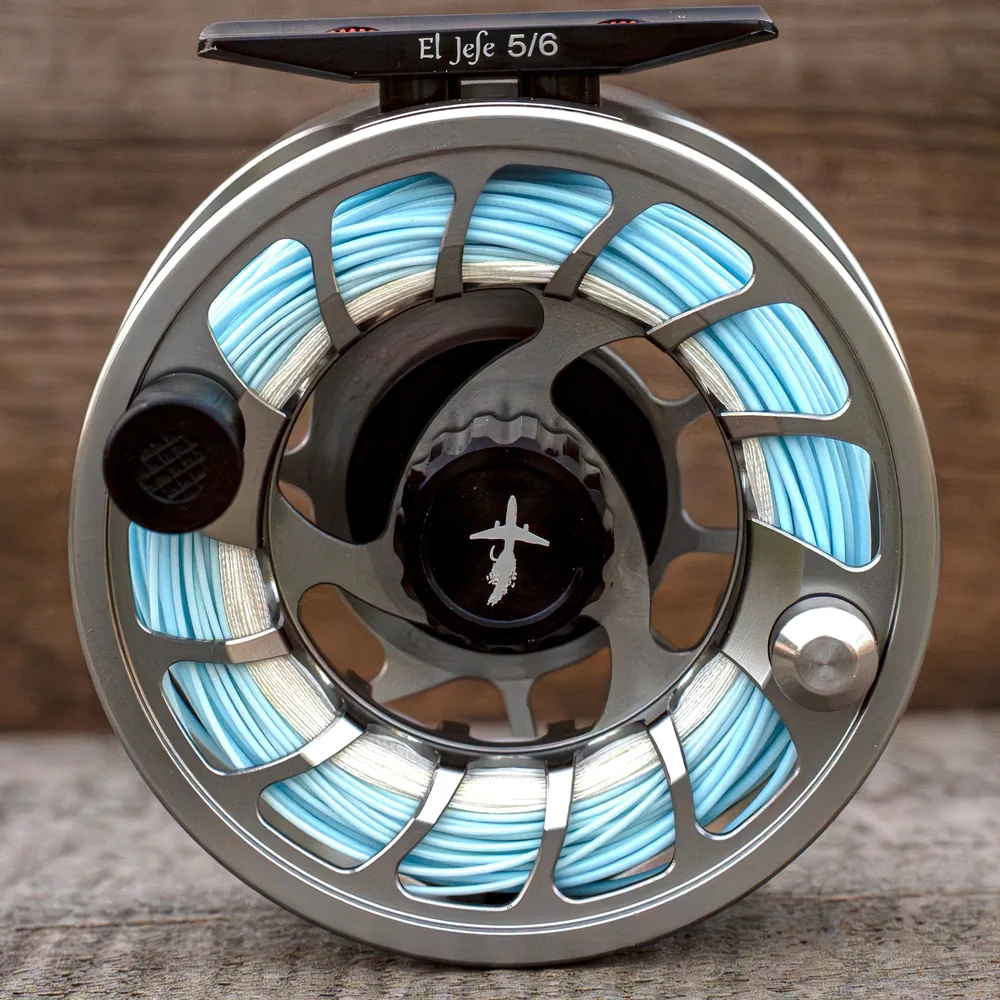 Face on view of the El Jefe 5/6 reel against weathered wood, El Jefe 5/6 printed on the black reel foot, the machined gunmetal frame around a spool already filled with pale blue fly line over white backing, black drag knob with the Pescador airplane mark at the center.